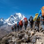 Eine Gruppe von Wanderern wandert unter klarem blauen Himmel einen schroffen Bergpfad hinauf, in Richtung schneebedeckter Gipfel. Die Bergsteiger sind mit Rucksäcken und Trekkingstöcken ausgestattet und bewegen sich durch eine atemberaubende