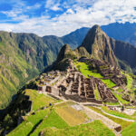 Weitwinkelaufnahme einer Bergkette in den Peruanischen Anden mit Wolken am blauen Himmel. Im Vordergrund in der Bildmitte die historischen Überreste des Machu Picchu einer alten Inkasiedlung auf einem Hochplateuau in ca. 2430m ü.N. Für ungeübte Reisende oder Reisende mit Vorerkrankungen kann eine Reisemedizinische Vorsorgeuntersuchung sinnvoll sein.