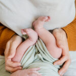 Gezeigt wird ein Neugeborenes Baby das von der Mutter oder dem Vater fest am Po gehalten wird.