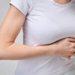 Gezeigt wird eine Frau die sich Schmerz geplagt den linken Rippenbogen Rand unterhalb der Brust hält. Das Bild möchte verdeutlichen was Schmerzen durch interkostalneuralgien auslösen.