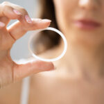 Das Bild zeigt einen Vaginalring zur Empfängnisverhütung in Großaufnahme, dahinter schemenhaft eine Frau.