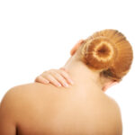 Gezeigt wird eine Frau mit nacktem Schulterrückengürtel die sich schmerzgeplagt an die oberen linken Schulterpartien greift. Gezeigt werden soll eine muskuläre Verspannung und Nacken- / Kopfschmerz.