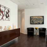 Das Bild zeigt unser Wartezimmer, Mit Fernseher, modernen Stühlen und einem Metallkunstwerk des israelischen Künstlers "JoJo" über einem Sideboard an der linken Wand.