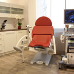 Das Bild zeigt ein Untersuchungszimmer mit einerseits einem gynäkologischen Behandlungsstuhl, links davon ein Schrank mit Schubladen und rechts ein Ultraschallgerät. Auf diesem symbolisch dargestellt ein Kind im Mutterleib.