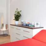 Das Bild zeigt eines unserer Behandlungs- und Untersuchungszimmer im Vordergrund eine der Behandlungsligen. Unter angeordnet ein Sideboard mit Schubladen.