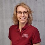 Manuela Franke, Blondhaarige Frau mit Brille in einem roten Shirt mit Aufschrift Frauengesundheit vor einer grauen Wand