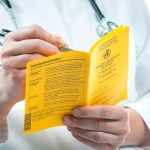 Arzt hält gelben Impfausweis in den Händen, trägt etwas handschriftlich ein